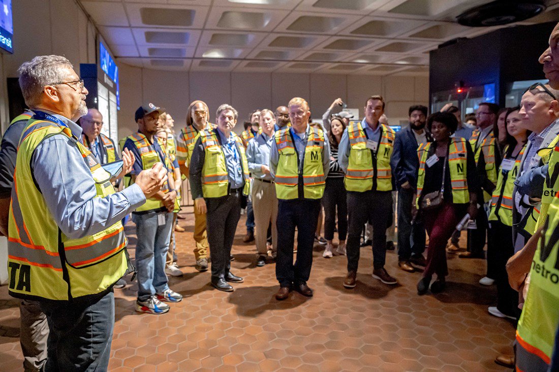 Tour participants in yellow safety vests listen to a metro representative talk at the entrance to a metro station.